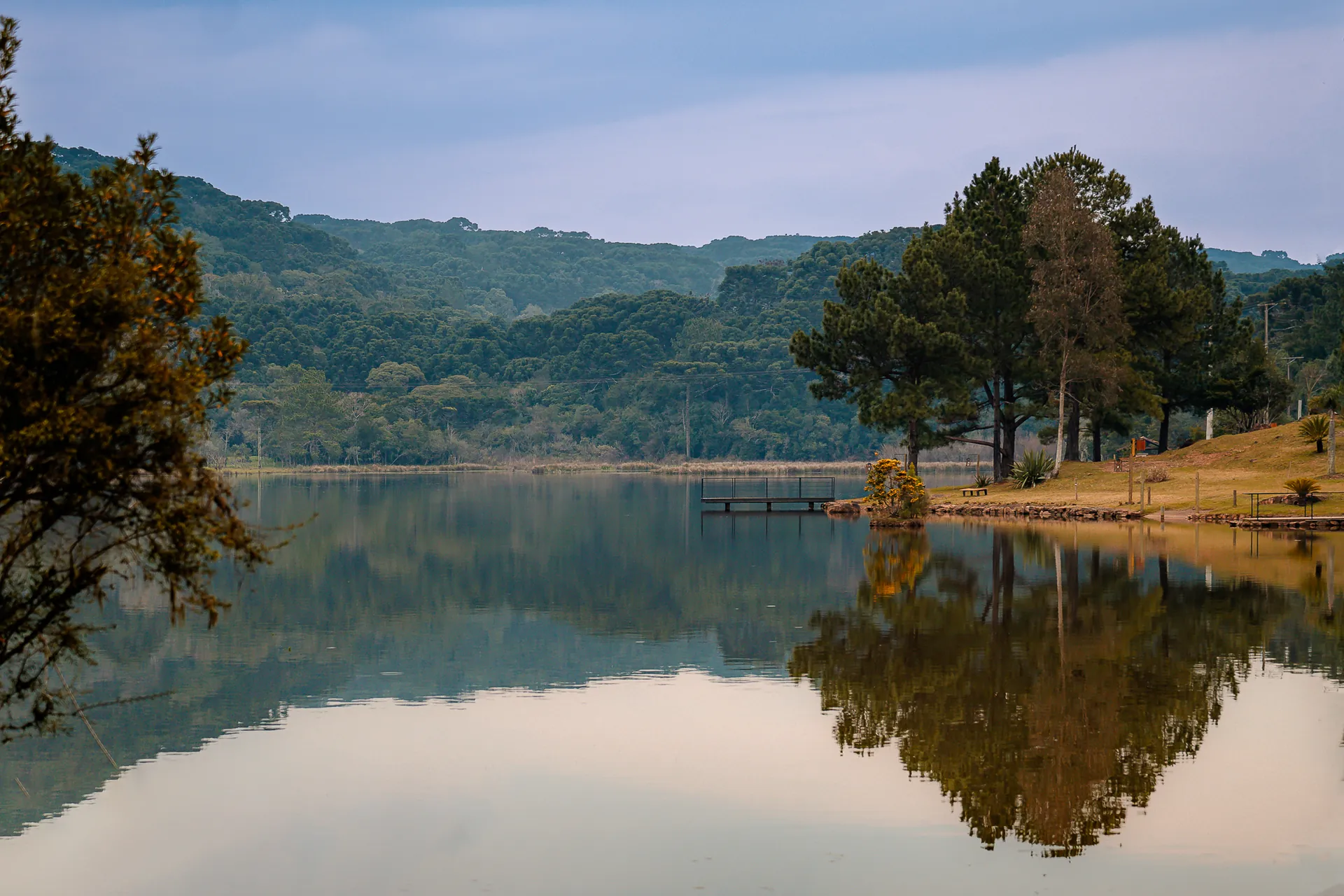 Lago de águas calmas refletindo a mata de araucárias e as colinas, com um píer de metal e a margem gramada
