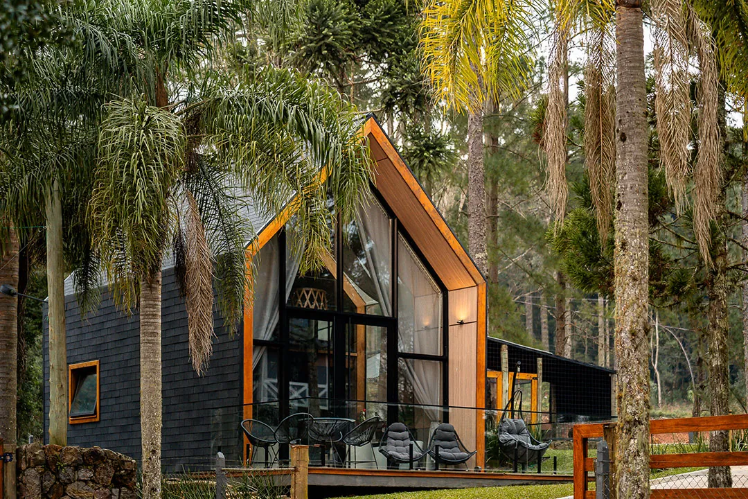 Lake house de fachada envidraçada com moldura de madeira e deck com cadeiras, entre palmeiras e araucárias