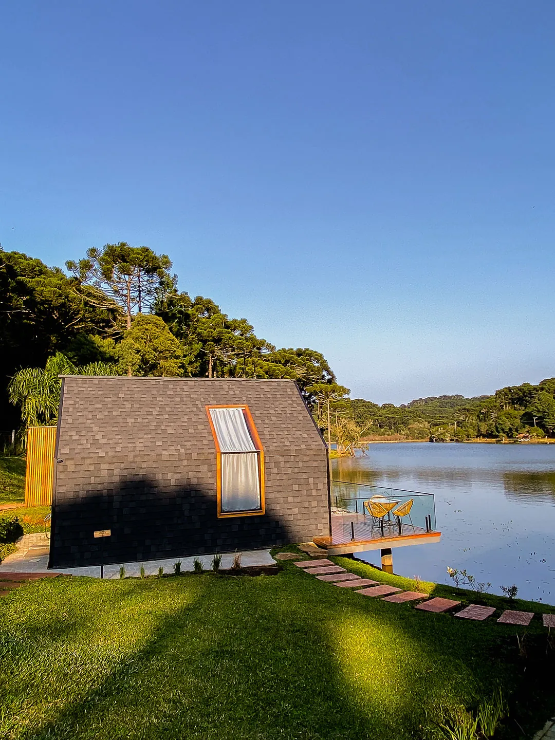 Lake house de telhas escuras com janela saliente e deck de guarda-corpo de vidro sobre o lago, gramado com caminho de pedras e céu limpo