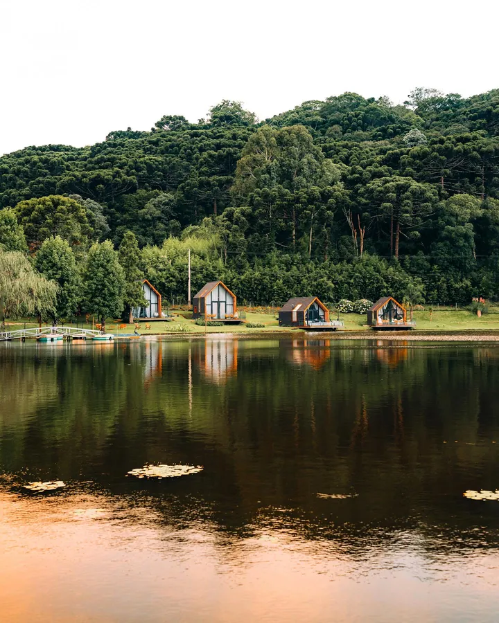 Lago com reflexo da mata e quatro lake houses enfileiradas na margem oposta, vitórias-régias na água e encosta coberta de araucárias.