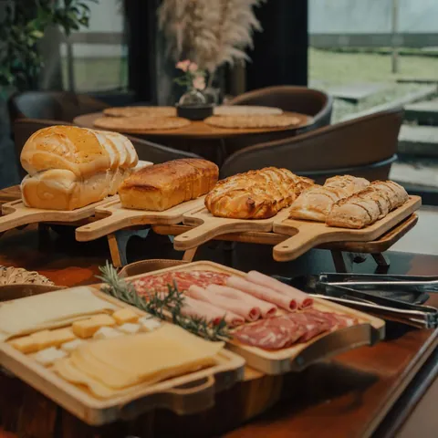 Pães inteiros e fatiados sobre tábuas de madeira elevadas e travessas com queijos, presunto e salame, com mesa redonda e poltronas ao fundo.