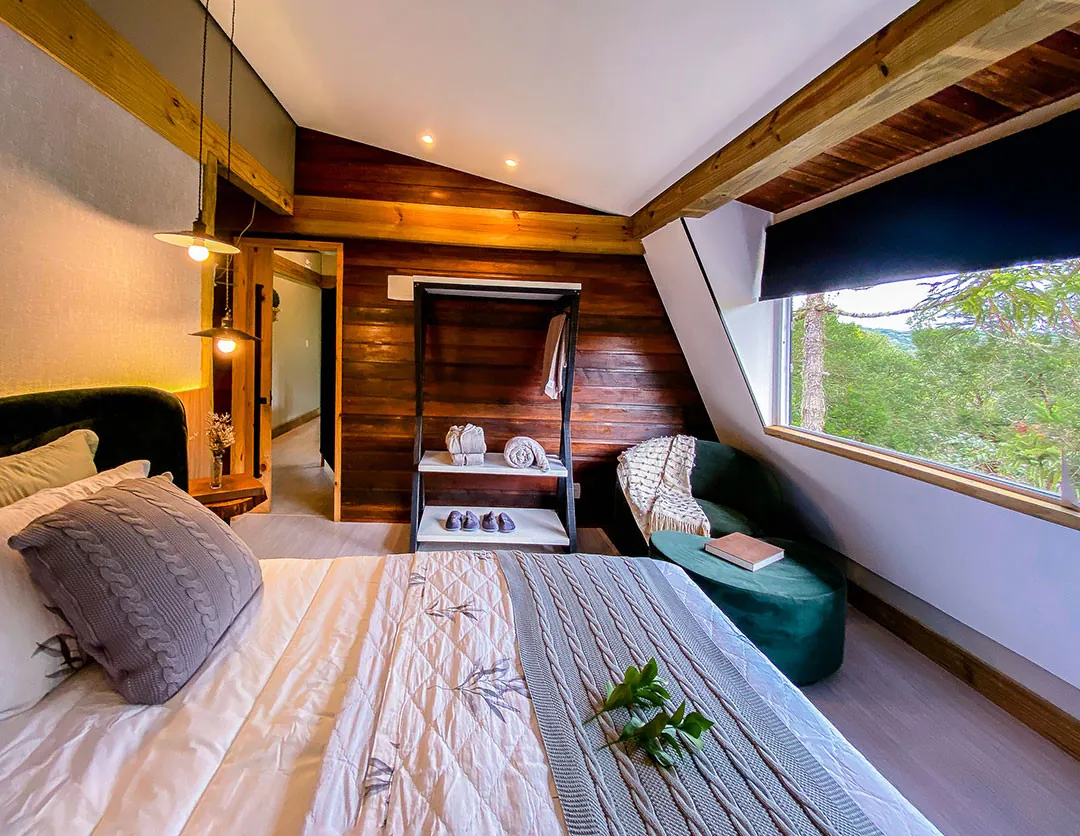 Quarto de teto inclinado com cama em primeiro plano, almofada de tricô cinza e ramo verde; ao fundo, parede de tábuas de madeira, estante com toalhas e pantufas, poltrona verde e janela ampla.