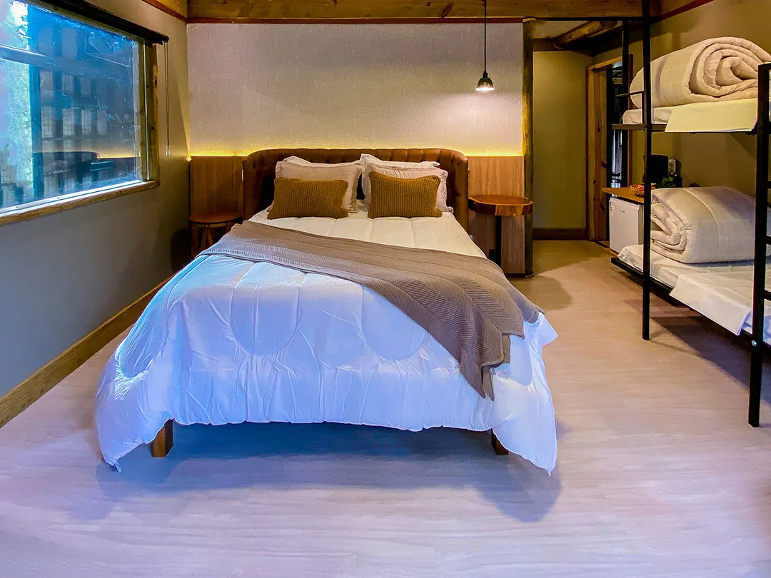 Cama de casal com edredom branco e manta marrom, cabeceira estofada e iluminação indireta; à direita, beliche com colchões e cobertores; janela à esquerda.